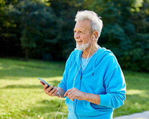 Happy active senior man having fun using smartphone and wearing earphones and sportswear, checking music or an exercise app, after having an exercise sport activity outdoors