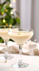 Two creamy cocktails in coupe glasses sit on a white surface with a blurred background of lights and greenery