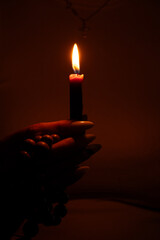 Woman's hand holding a lit candle and wooden rosary beads in the dark at home, flame illuminates fingers, faith