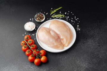 Raw chicken fillets on white plate with salt, tomatoes and herbs on black table