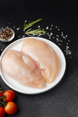 Raw chicken fillets on white plate with salt, tomatoes and herbs on black table