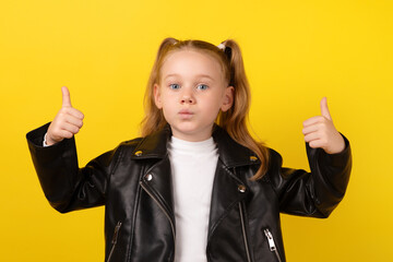 Young girl in a black leather jacket making a surprised face and showing thumbs up against a yellow background.