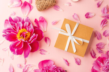 Gift box with white ribbon surrounded by peonies, petals, candles and twine on pink background