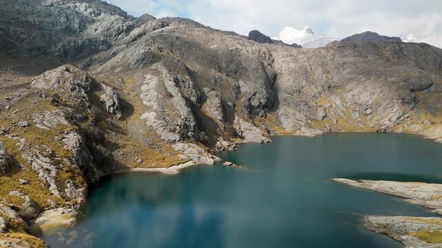 A stunning aerial flyover reveals a pristine, deep blue alpine lake at Paso del Zorro, nestled among the rugged, rocky peaks of the remote Huayhuash mountain range in the Peruvian Andes.