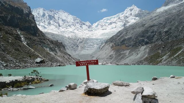 A first-person point-of-view shot shows a tourist hiking past the stunning, turquoise glacial waters of Laguna Llaca at the base of snow-capped peaks in Peru's Huascar&aacute;n National Park.