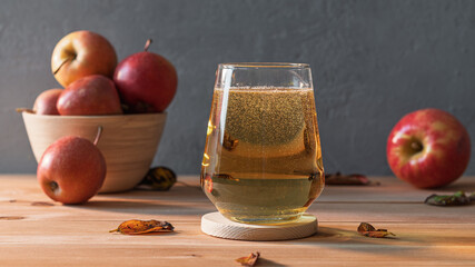Sparkling golden apple cider in modern glass on wooden coaster, fresh apples in background add natural autumn harvest charm.