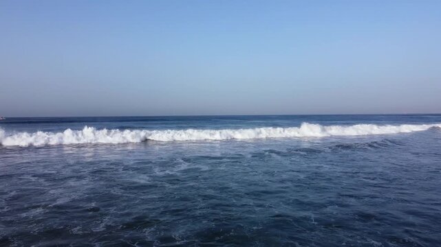 Aerial drone footage starting at the shore and moving over the sea, with a breaking wave, two swimmers, and a clear sunny sky.
