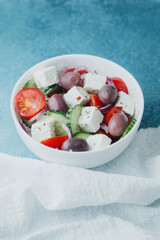 Greek salad with feta cheese olives cucumbers tomatoes and onions in white bowl on blue background