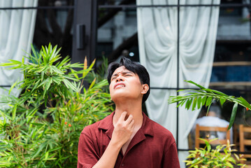 A distressed young asian man rubbing his neck with a pained expression, showing signs of a sore throat and unease during a quiet moment at a cafe.
