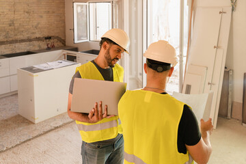 Construction workers discussing plans using laptop in an unfinished kitchen during house renovation