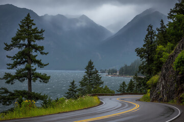 A winding road curves alongside a lake surrounded by evergreen trees and misty mountains under a cloudy, overcast sky