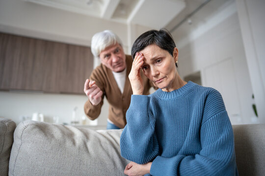 Distressed mature wife hiding from yelling husband, enduring aggression from neurotic man in mental instability. Caregiver stress, manipulative control, suppressed emotions, senior relationship crisis