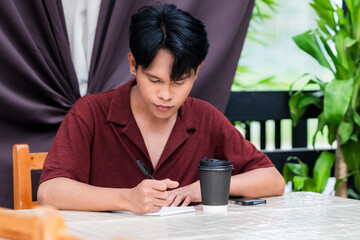 A focused young asian man deeply engrossed in writing on his notepad, pensive and thoughtful as he...