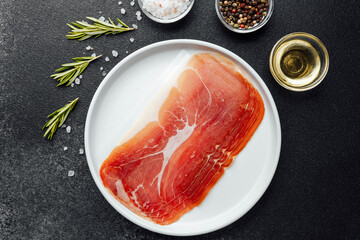Sliced prosciutto ham on white plate with herbs and spices on dark background