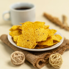 White plate filled with crispy yellow tortilla chips, placed on a piece of burlap cloth, accompanied by a cup of black coffee and green plant decoration on a light background.
