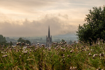 Fototapeta premium Sunrise over misty hills with a church steeple rising above wildflower fields
