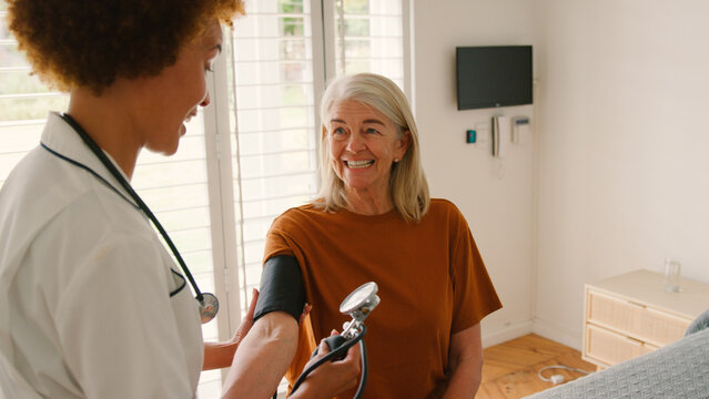 Female Nurse Wearing Uniform Taking Blood Pressure Of Senior Woman In Private Hospital Room