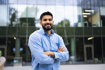 A smiling Indian man stands confidently with arms crossed in front of a modern glass building.