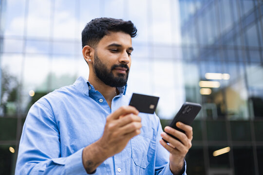 An Indian man in a blue shirt is shown holding a credit card and a smartphone in front of a modern building.