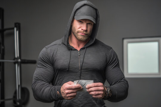 Muscular man in a dark hoodie and cap stands indoors, holding a folded banknote, with gym equipment and a window in the background