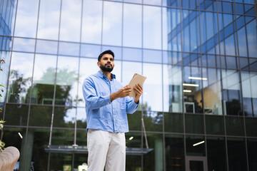 A man in a blue shirt stands in front of a modern glass building, holding a tablet, looking up.