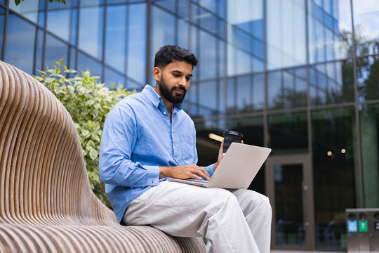 A man sits on a bench, using a laptop outdoors, with a coffee cup in hand, in front of a modern building. - Powered by Adobe