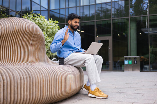 A man sits outdoors, working on a laptop while waving. He is smiling and appears to be in a video call, enjoying a coffee break.