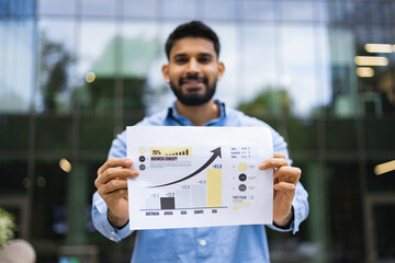 A smiling man holds a chart showing business growth outdoors with a blurred building in the...