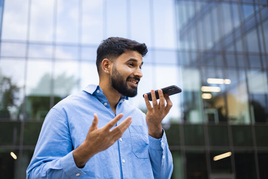 An Indian man is using voice command on his smartphone, smiling, and gesturing in front of a modern building.