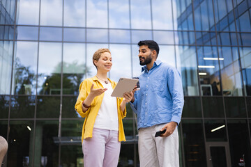 Two business colleagues discuss on a tablet outdoors in front of a modern office building.