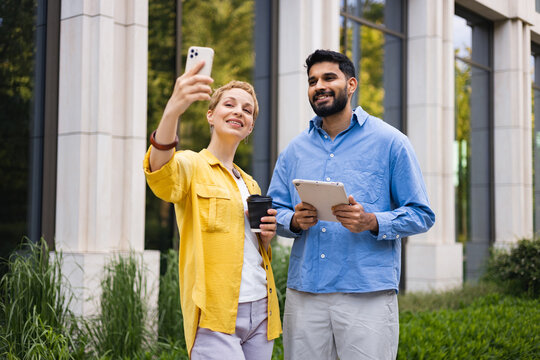 A diverse couple taking a selfie outdoors with a smartphone and tablet in front of a modern building.