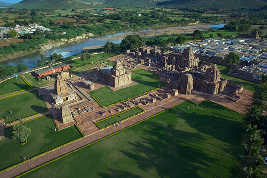 Aerial view of ancient temples standing majestically amidst lush green lawns, bordered by a serene river and quaint village homes, Badami, Karnataka, India.