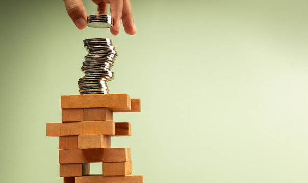 Hand stacks coins atop unstable Jenga tower, symbolizing high risk investments and volatile economic conditions - Powered by Adobe
