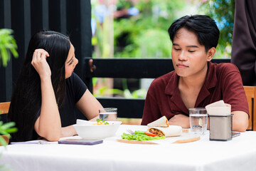 A young Asian man looking down disappointed while a young Asian woman watches him closely, both caught in a subtle couple quarrel filled with tension and unspoken feelings at a cafe