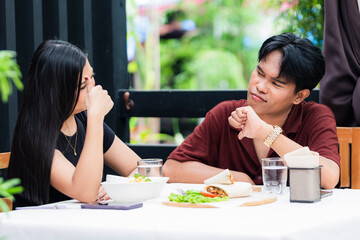 A young Asian man showing a thumbs down while a young Asian woman confidently displays a thumbs up, capturing a playful yet clear moment of couple disagreement and contrasting opinions
