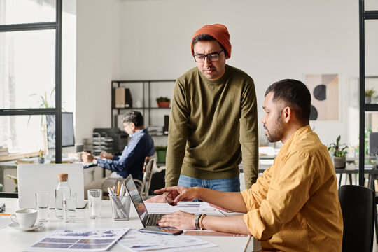 South Asian man sitting at desk in office showing presentation on laptop screen to his coworker, copy space