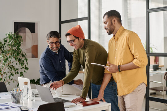 Medium long shot of three cheerful young and mature Indian men working on business project in modern office
