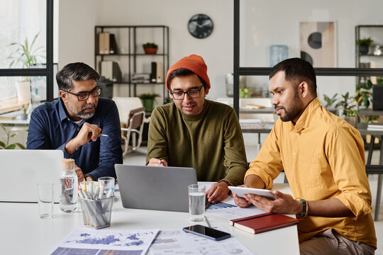 Group of three Indian men sitting at table in modern office working on business project, looking at laptop screen and discussing plan
