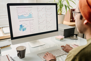 Selective focus shot of young specialist sitting at table in modern office working on charts and graphs on desktop computer