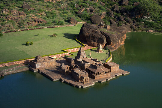 Aerial view of the ancient temple complex reflected on the tranquil waters of the lake, surrounded by rugged cliffs and verdant meadows, Badami, Karnataka, India.