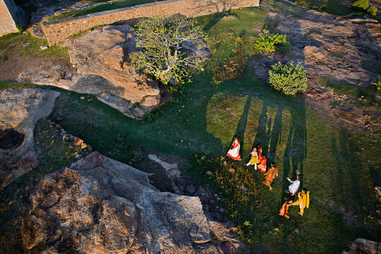 Aerial view of women in vibrant saris gather on the grassy rocks, casting long shadows in the soft afternoon light, Madurai, Tamil Nadu, India.