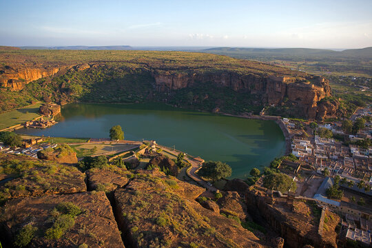 Aerial view of a serene lake embraced by rugged cliffs and verdant trees, contrasting with the clustered buildings, Badami, Karnataka, India.