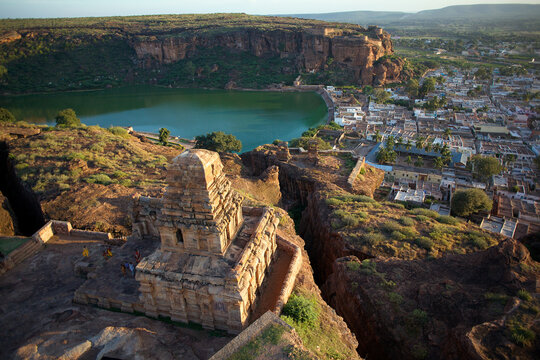 Aerial view of ancient stone temples perched on rugged cliffs overlooking a serene lake and town, Badami, Karnataka, India.