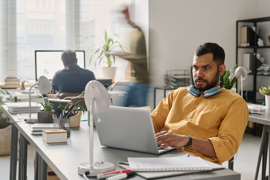 Young South Asian man wearing yellow shirt sitting at desk in modern office working on laptop, copy space