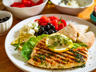 Greek cuisine: grilled chicken breast with lemon, pita bread and greek salad on wooden table	