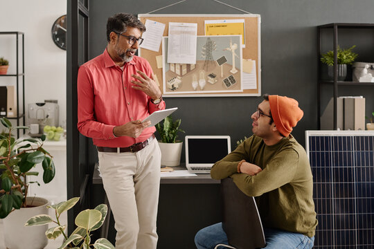 Medium long shot of mature Indian man holding digital tablet standing in office talking to his younger colleague