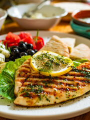Greek cuisine: grilled chicken breast with lemon, pita bread and greek salad on wooden table	