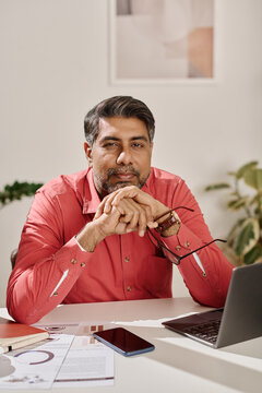 Vertical medium portrait of mature South Asian man wearing coral shirt sitting at desk in office looking at camera