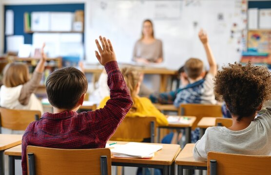 Diverse group of elementary school students raising hands to answer question during lesson, teacher standing at front of classroom