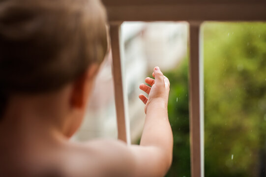 A young child stands indoors, looking out of a window on a rainy day. Their hand is pressed against the window, fingers outstretched as if trying to touch the falling raindrops. - Powered by Adobe